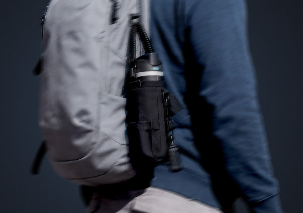 Showing person in navy jacket with hood up and carrying the Spigen tumbler holder bag with water bottle the pocket attached to backpack