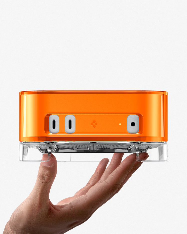 Hand holding Spigen Mac Mini Case Stand in orange from below showing front port cutouts and clear acrylic base
