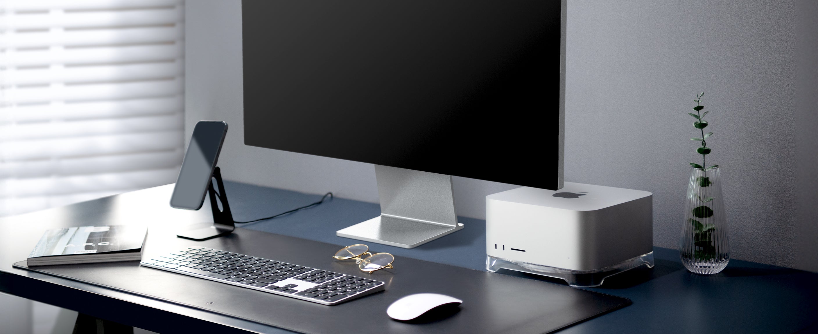 Desk showing Mac Mini, iMac, keyboard, mouse, glasses, iPhone on MagFit Stand, Spigen desk mat, magazine, and eucalyptus in vase