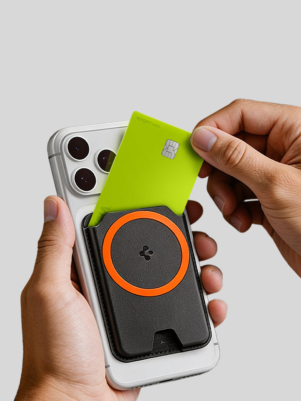 Hands pulling a card out of a Spigen MagSafe wallet in black with orange ring accent attached to the back of an iPhone