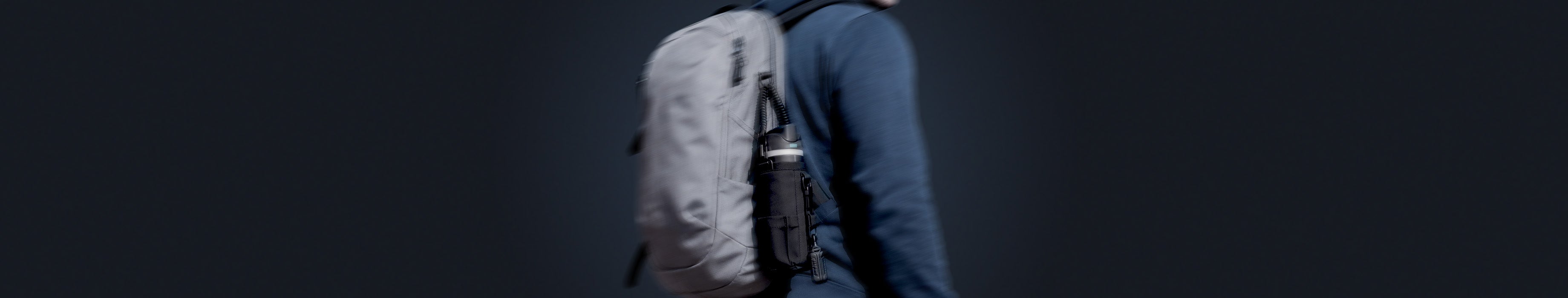 Showing person in navy jacket with hood up and carrying the Spigen tumbler holder bag with water bottle the pocket attached to backpack